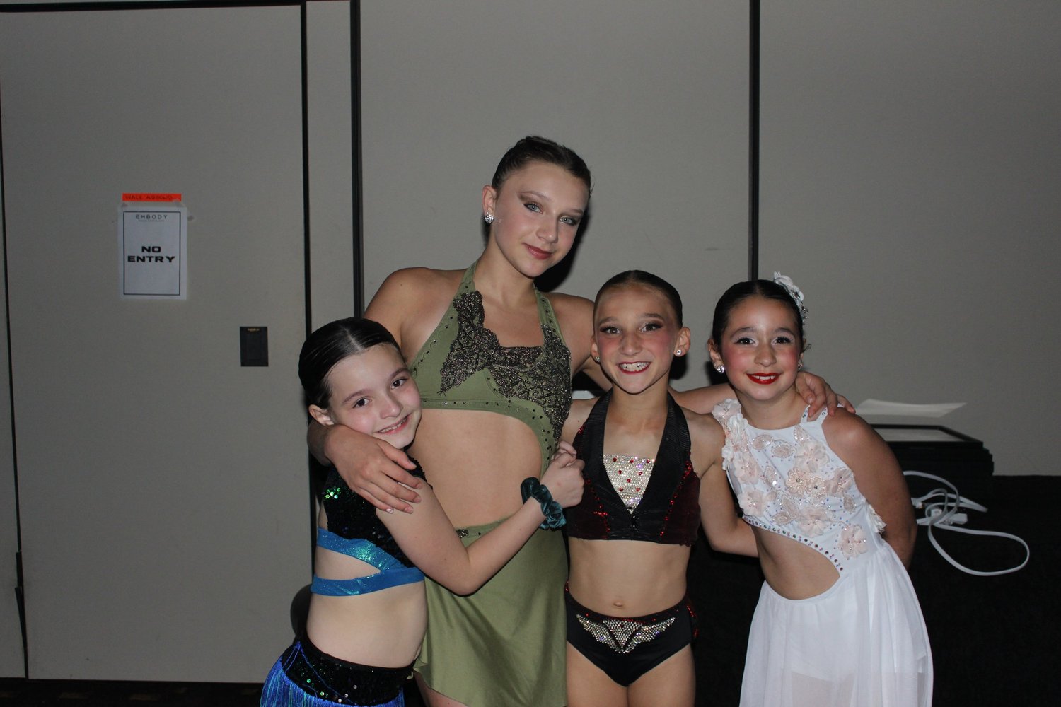 Four dancers in different competition costumes with their arms around one another.